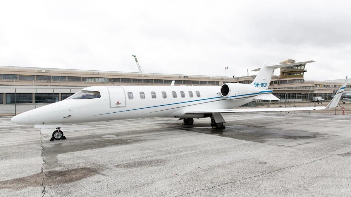 A white Learjet with a thin blue stripe parked on a wet tarmac in front of an airport terminal building.