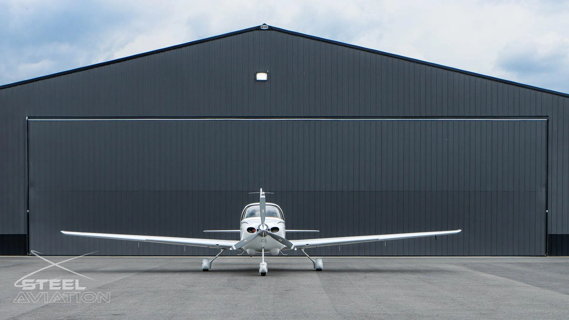 A white Cirrus SR22 aircraft is parked head-on in front of a large dark grey hangar.