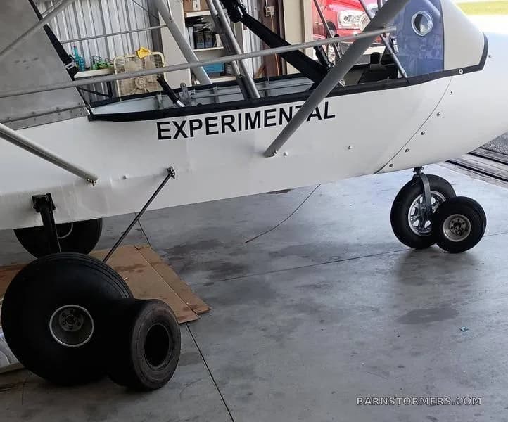 A close-up view of the white fuselage of an experimental ultralight aircraft, showing the landing gear and "EXPERIMENTAL" text on the side, with spare tires on the floor.