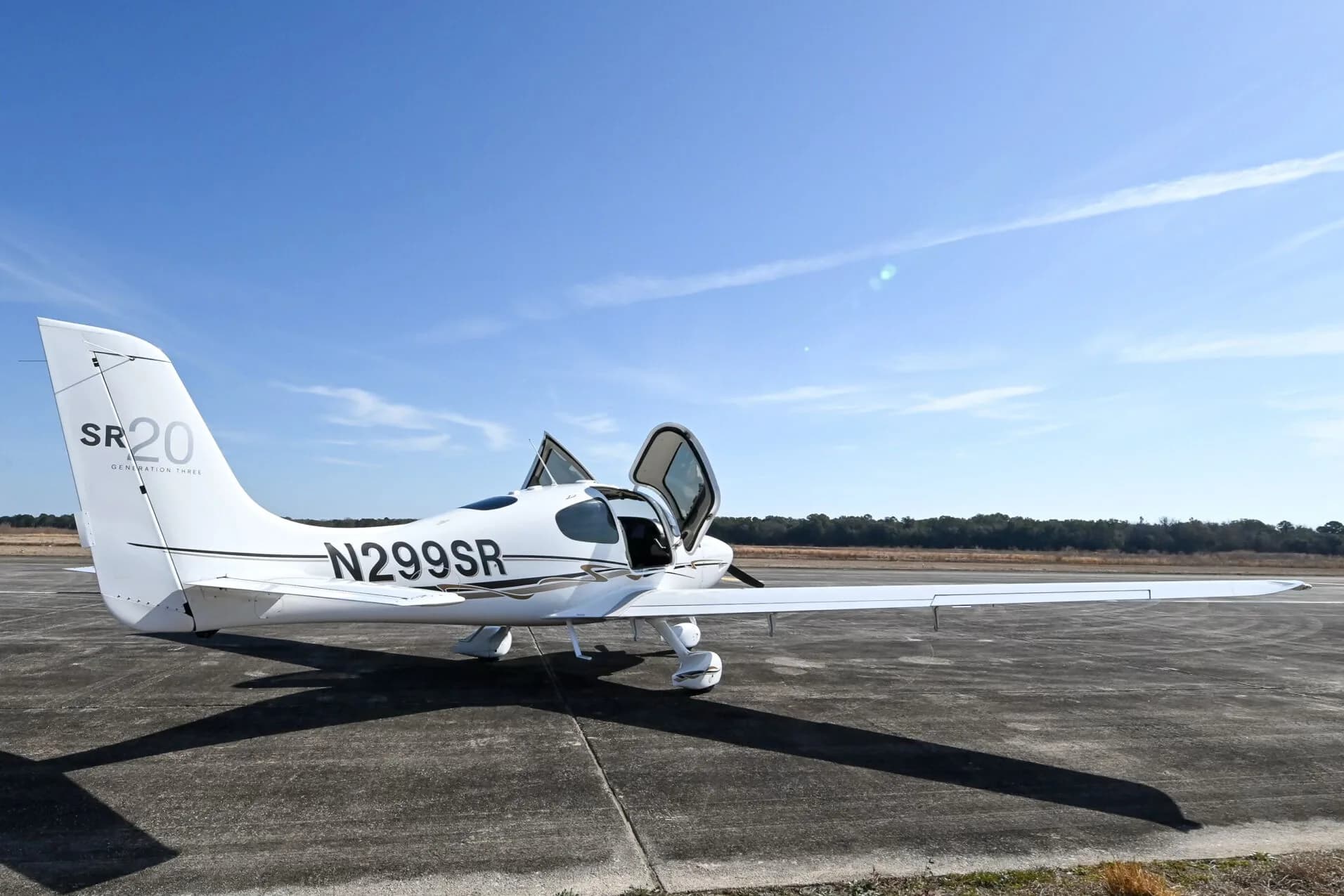 Rear-side view of a white Cirrus SR20 aircraft with black and gold accent stripes, registration N299SR, parked on an airfield with its doors open.