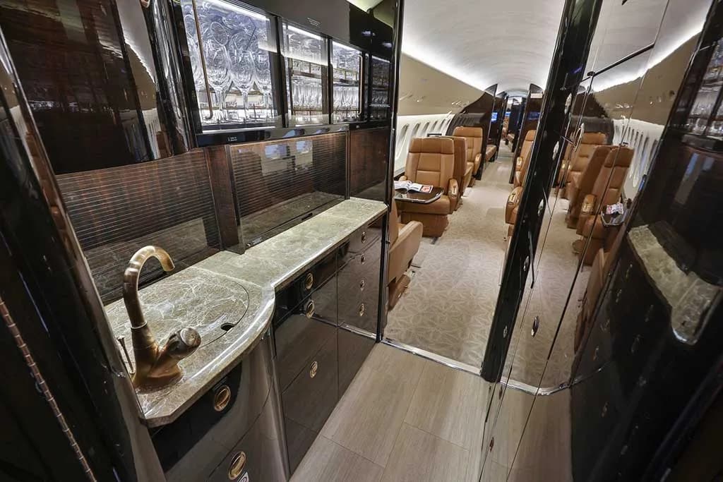 The aircraft's galley features a sink with a brass faucet, marble countertops, dark wood cabinetry, and a view into the main cabin.