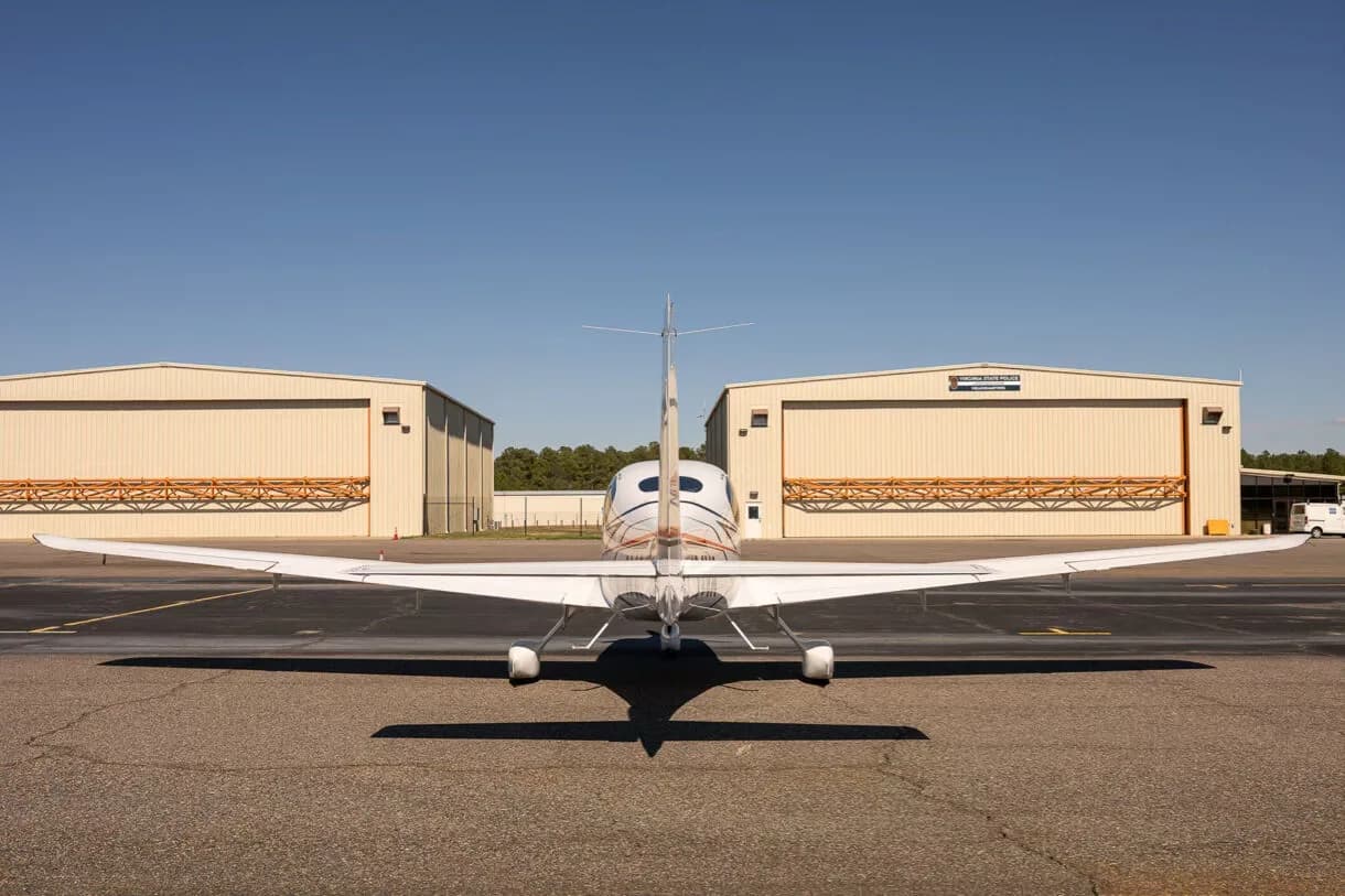 A rear view of a white Cirrus SR22 aircraft with hangars in the background, parked on an airfield.