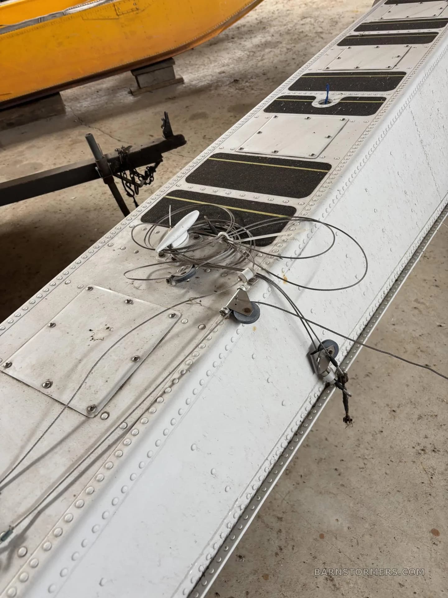 An overhead view of the top of a white aircraft float, detailing control cables, pulleys, and black anti-slip pads, with a yellow aircraft hull in the background.