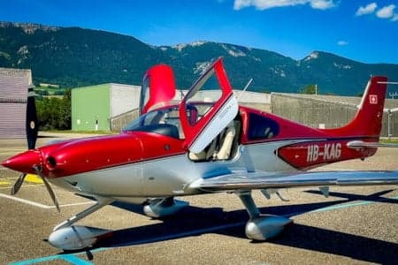 A red and silver Cirrus SR22 aircraft, registration HB-KAG, with open gull-wing doors, is parked on an airfield with mountains in the background.
