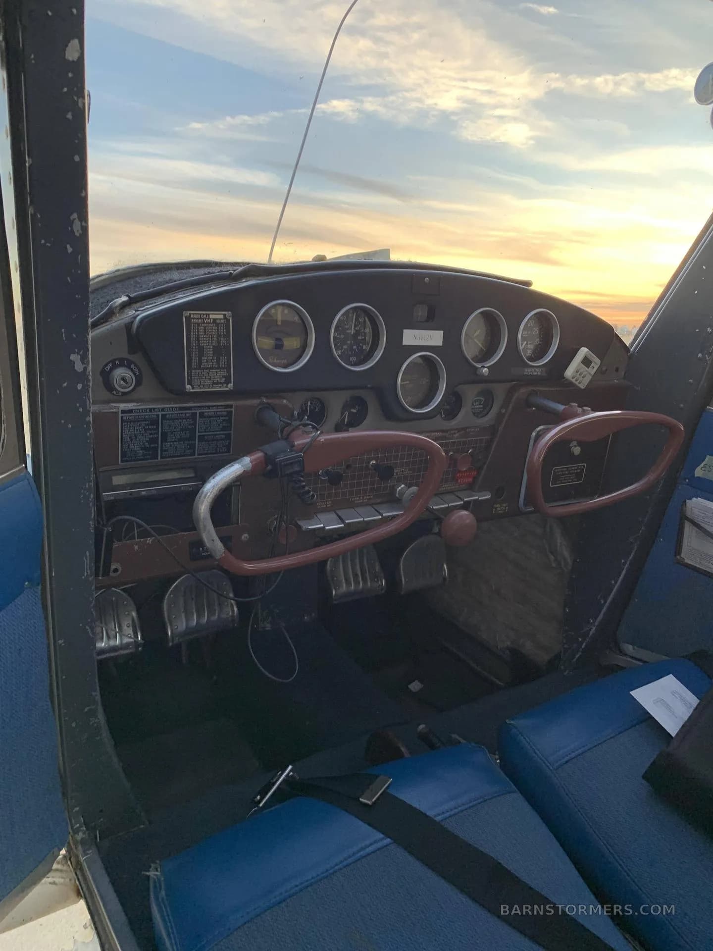 The cockpit of a Cessna 170B, showing the instrument panel and brown yokes, with a sunset sky visible through the windshield.