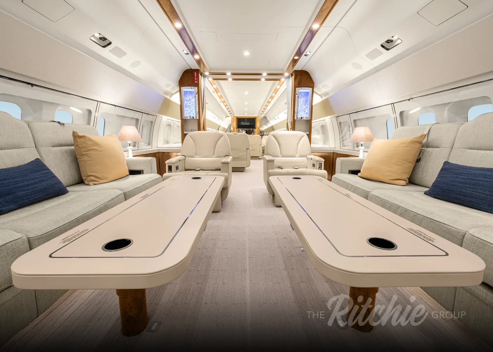 A view down the aisle of a luxurious aircraft cabin with light-colored sofas and tables.