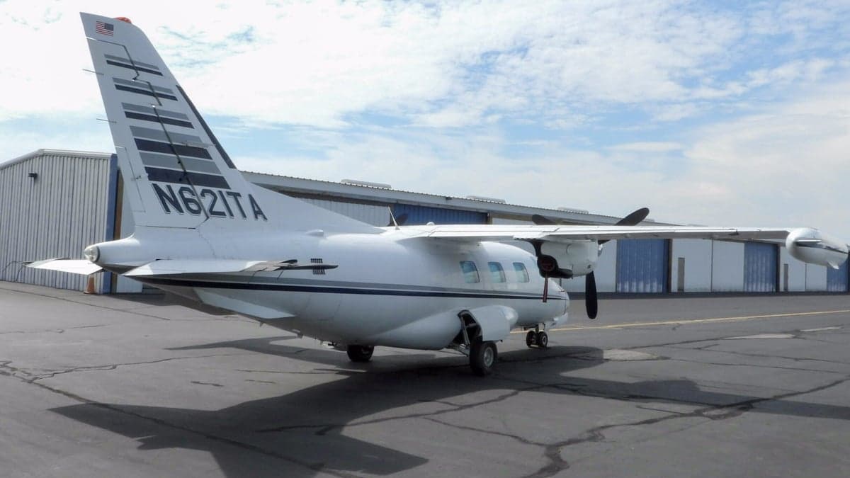 A white Mitsubishi MU-2 aircraft with black and grey stripes on the tail, parked on an asphalt ramp with hangars in the background.