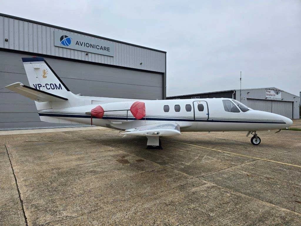 A white Cessna Citation jet, registration VP-COM, with red engine covers, parked in front of an Avionicaire hangar.