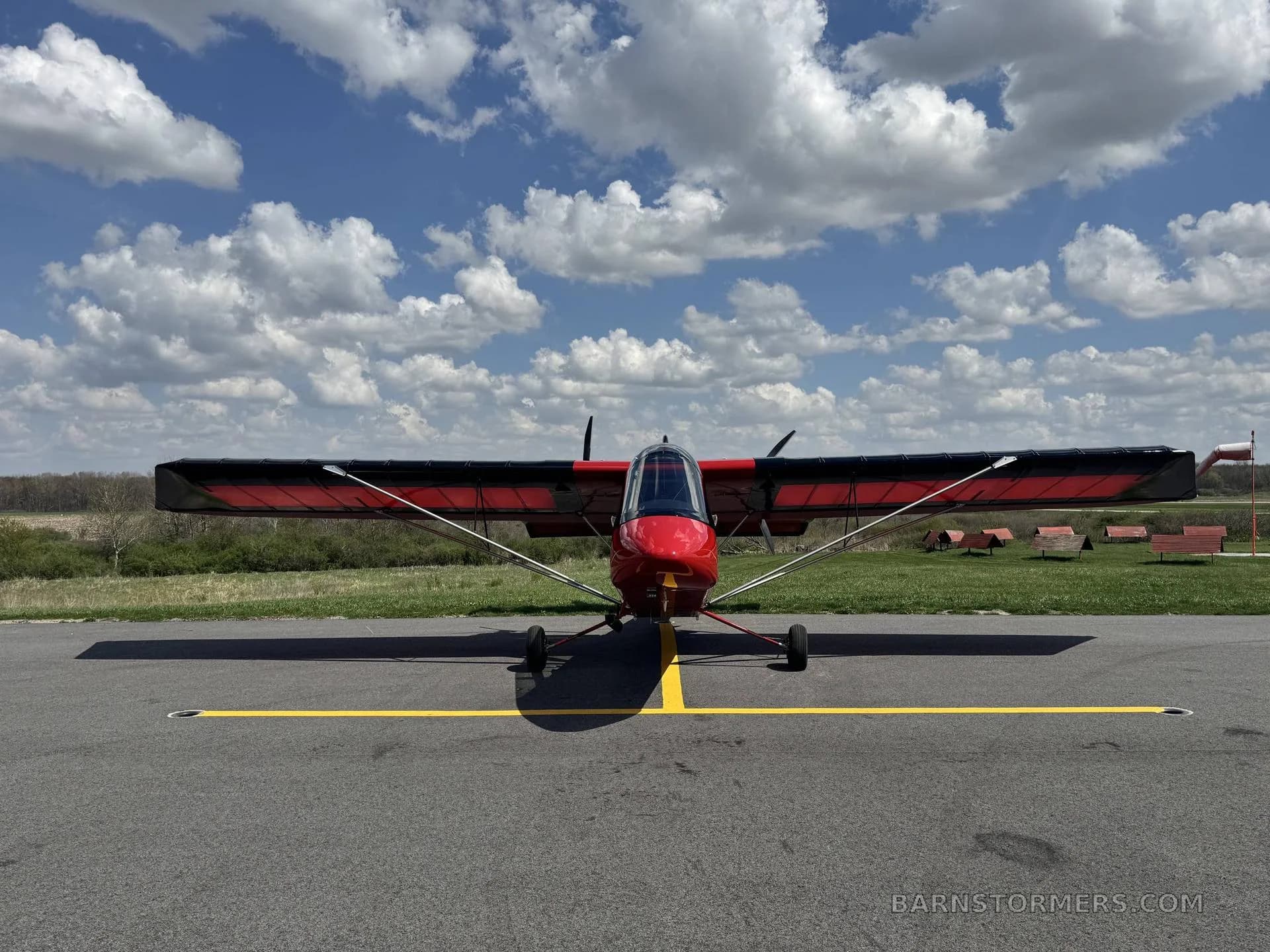 A red and black Air-Cam aircraft is parked head-on on an asphalt runway under a blue sky with white clouds.