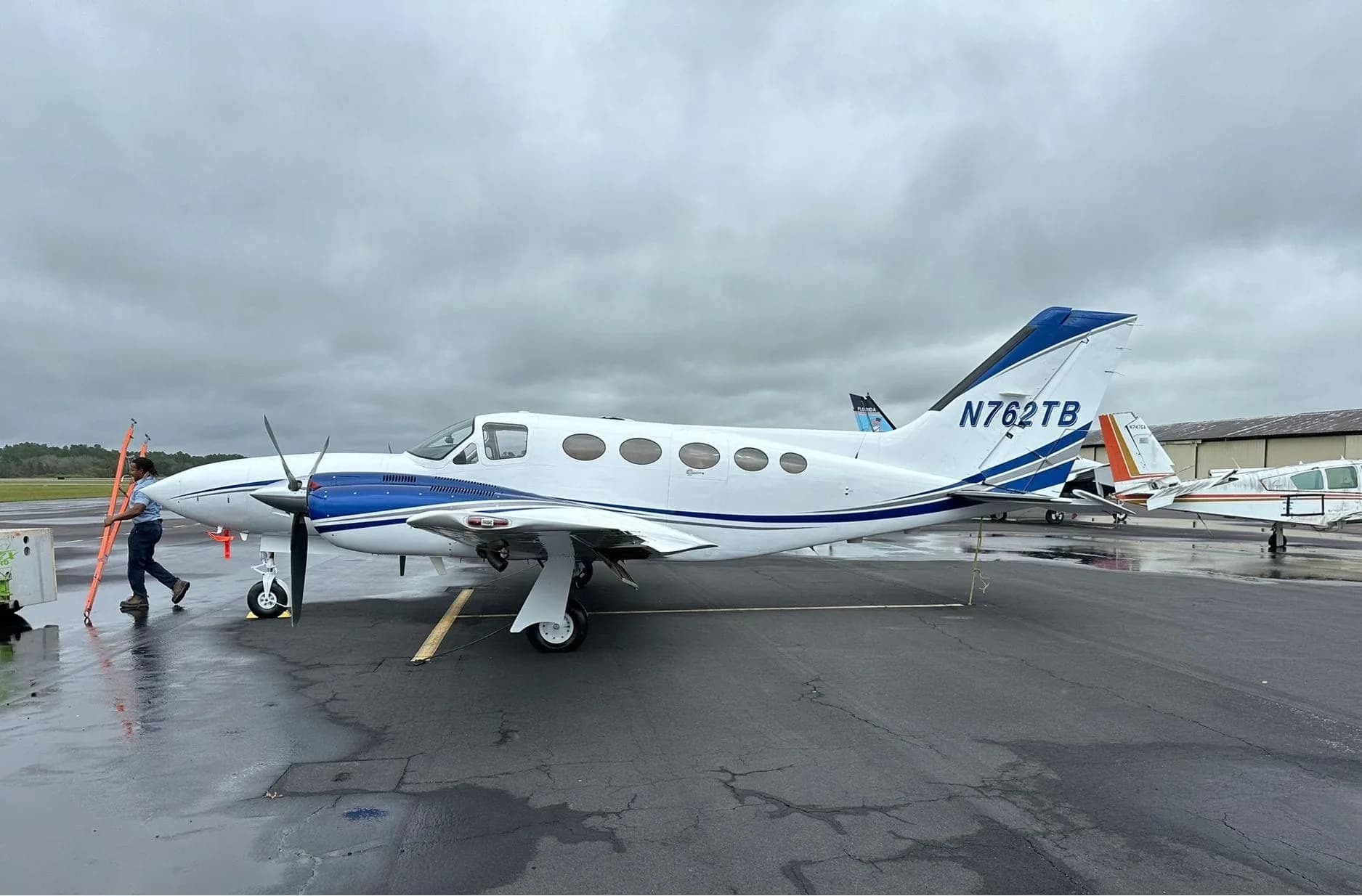 A white and blue twin-engine Cessna 414 Chancellor aircraft with registration N762TB parked on a wet tarmac under a cloudy sky.