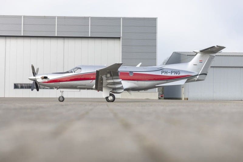 A Pilatus PC-12 aircraft with a silver and red paint scheme, registration PH-PNG, parked on the tarmac in front of a hangar.