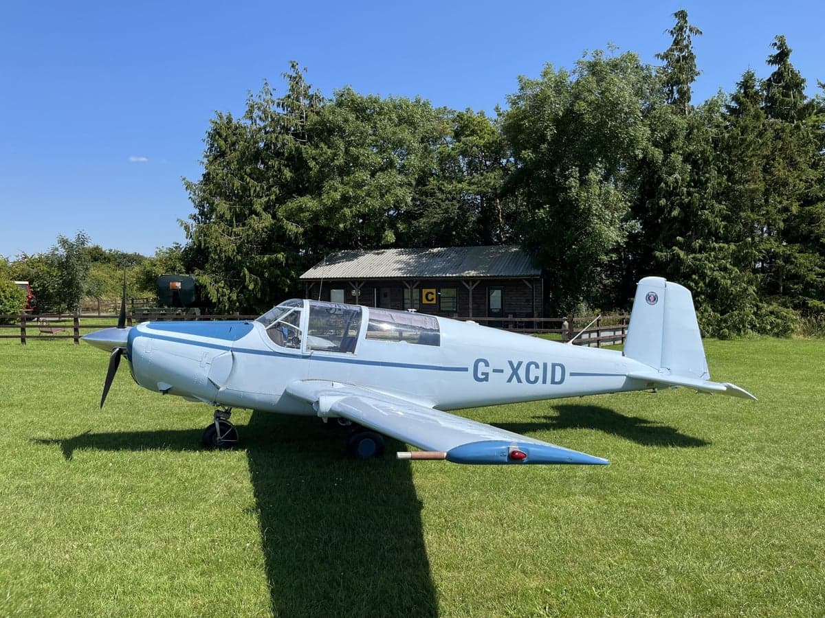 A light blue aircraft, registration G-XCID, sits on a grassy field.
