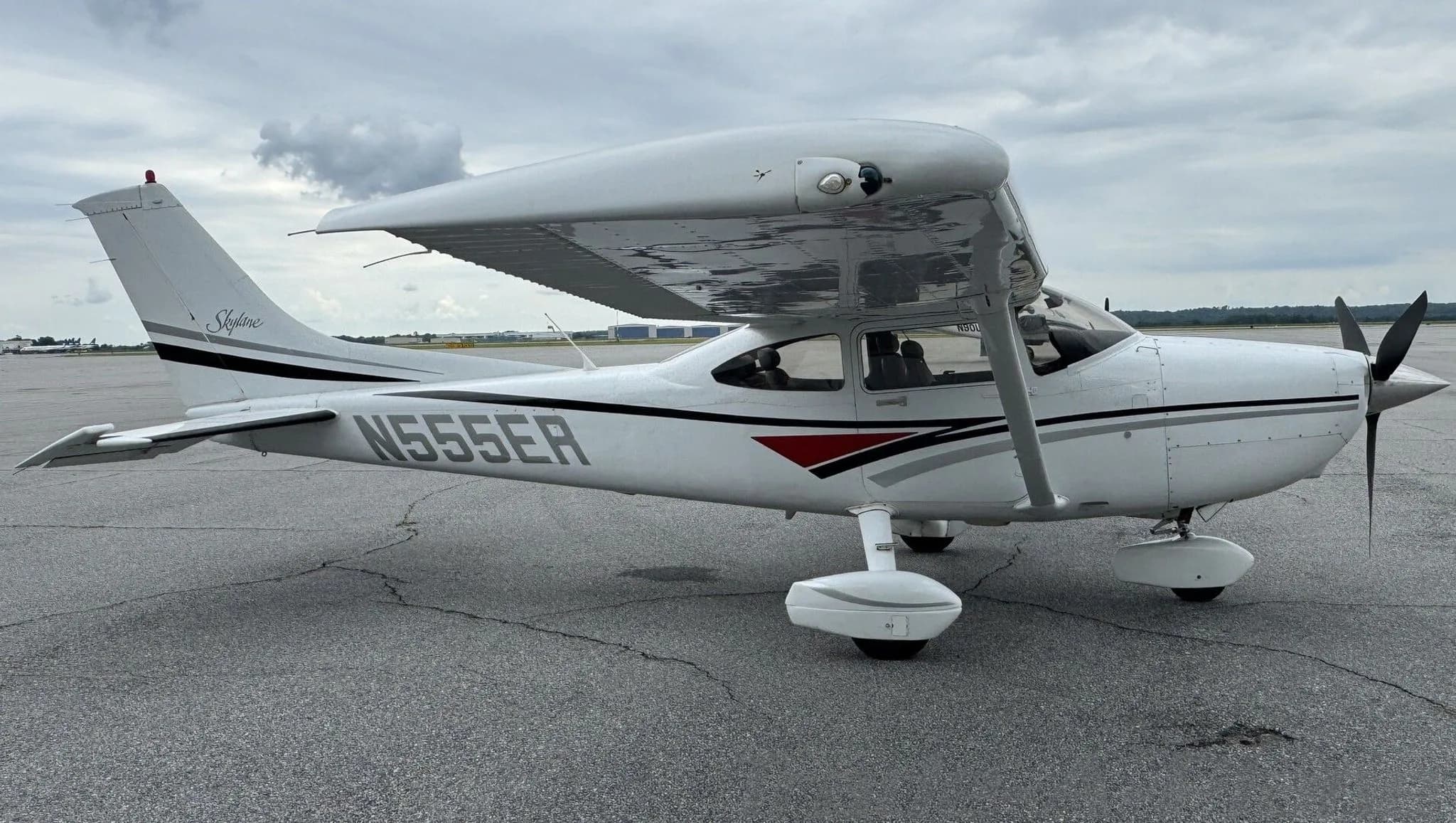 A white Cessna 182 Skylane with black, grey, and red stripes parked on an airfield under a cloudy sky.