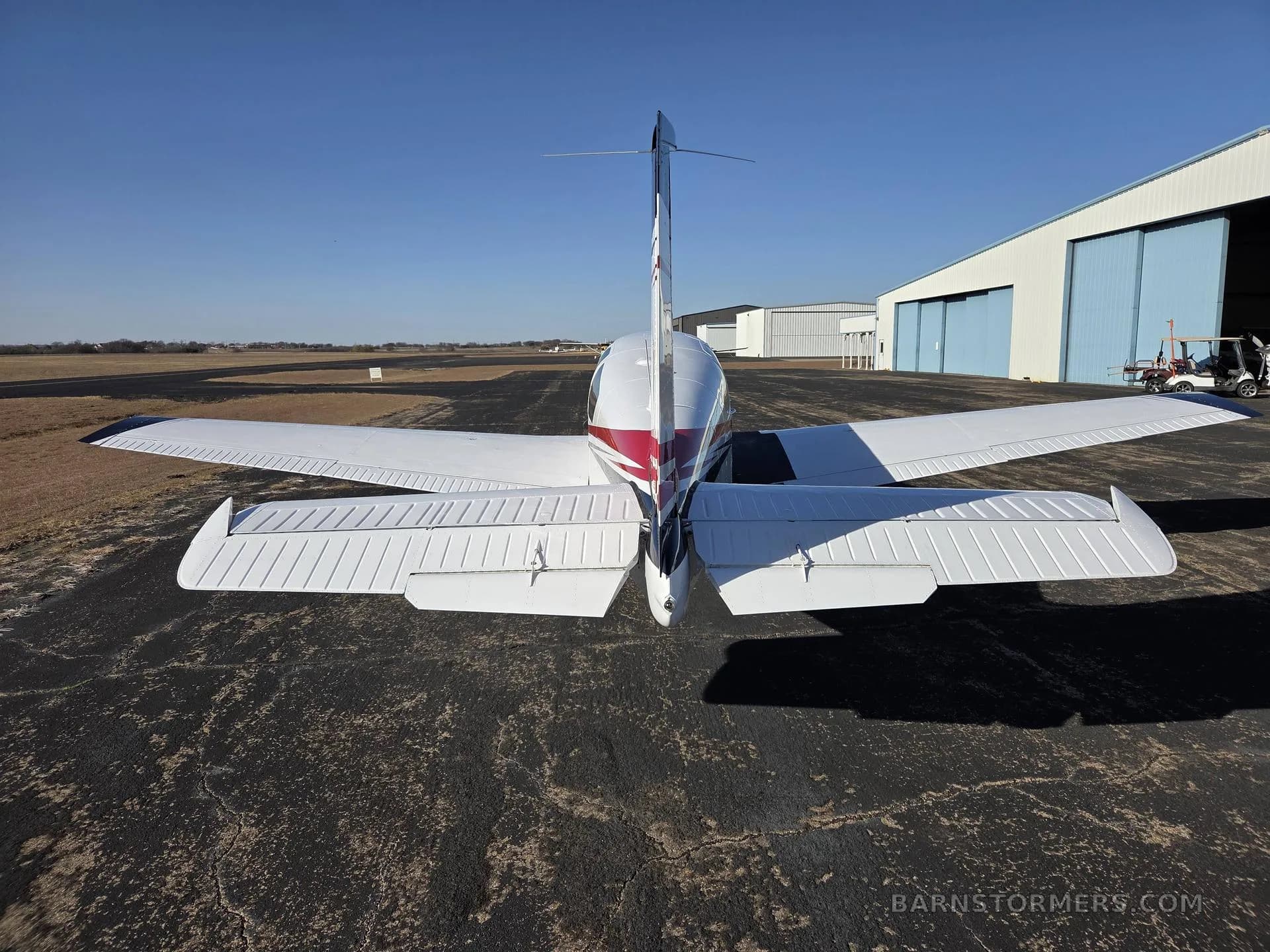 A rear view of a white single-engine aircraft with red and black stripes on the tail is parked on an asphalt tarmac near hangars.