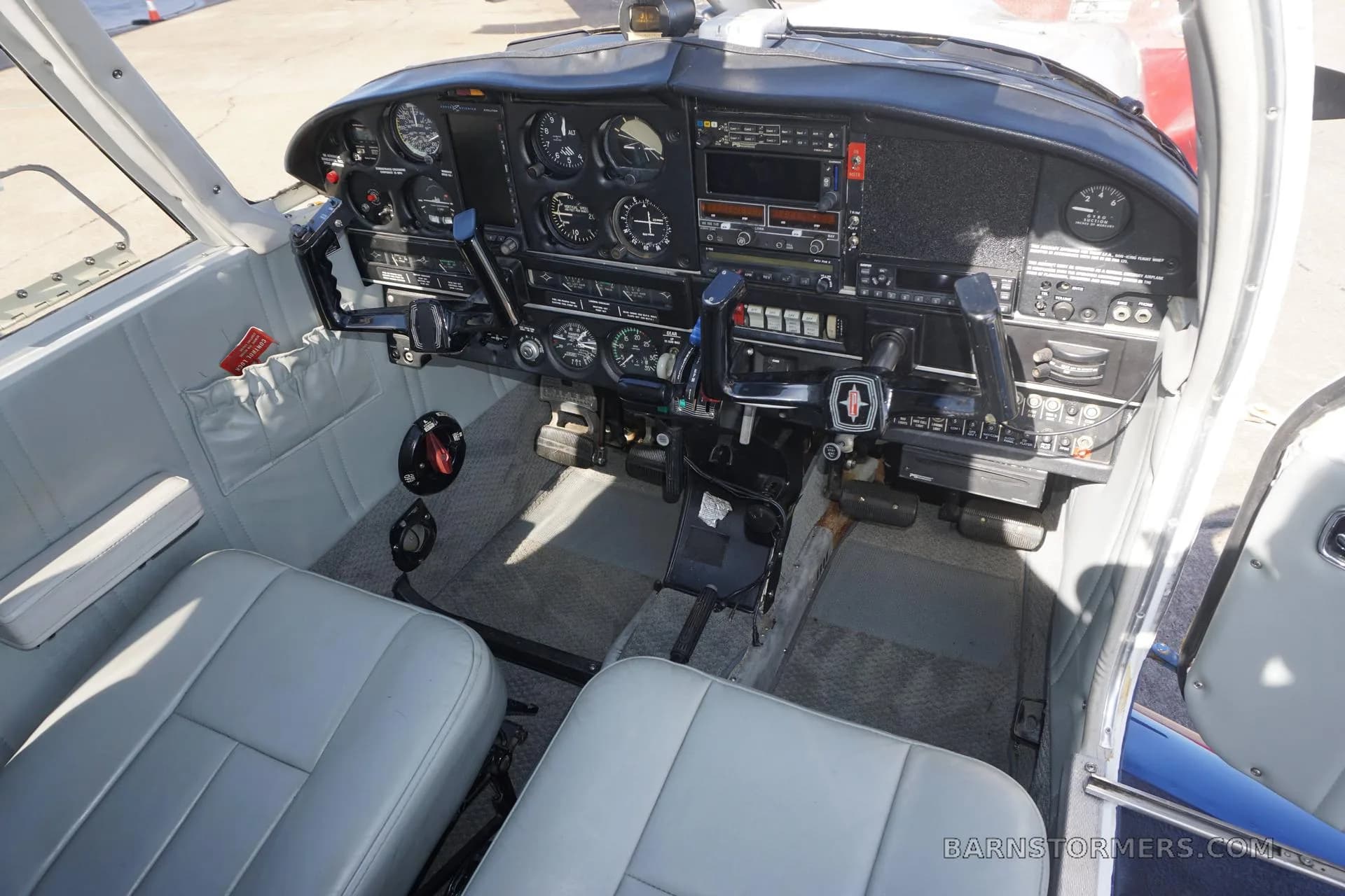 The cockpit of a small aircraft features a black instrument panel with various gauges and controls, and light gray seats.