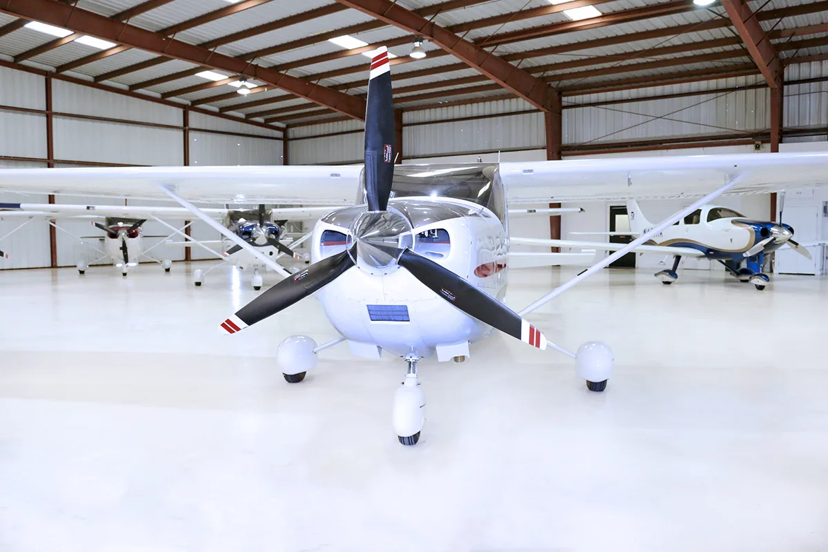 A front view of a white Cessna 182 Skylane with a three-bladed propeller, parked in a hangar.