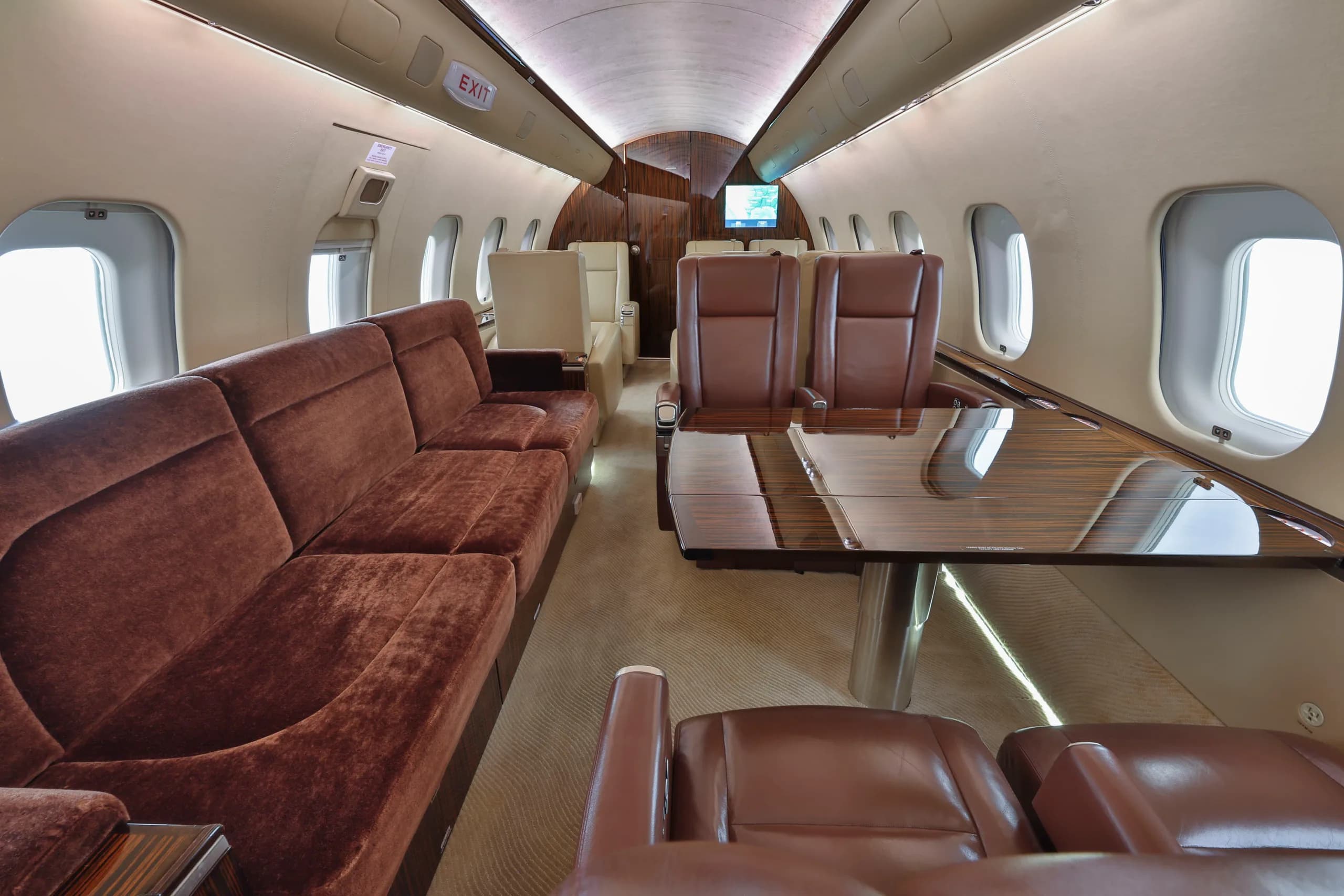 The rear section of the business jet's cabin, featuring a long brown velvet sofa and a dining area with a wooden table.