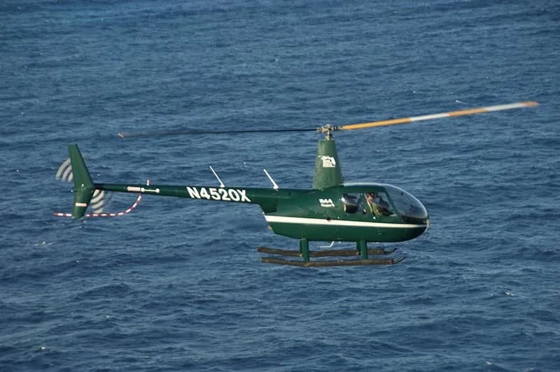 A dark green Robinson R44 Clipper II helicopter, registration N4520X, flies low over blue ocean water.