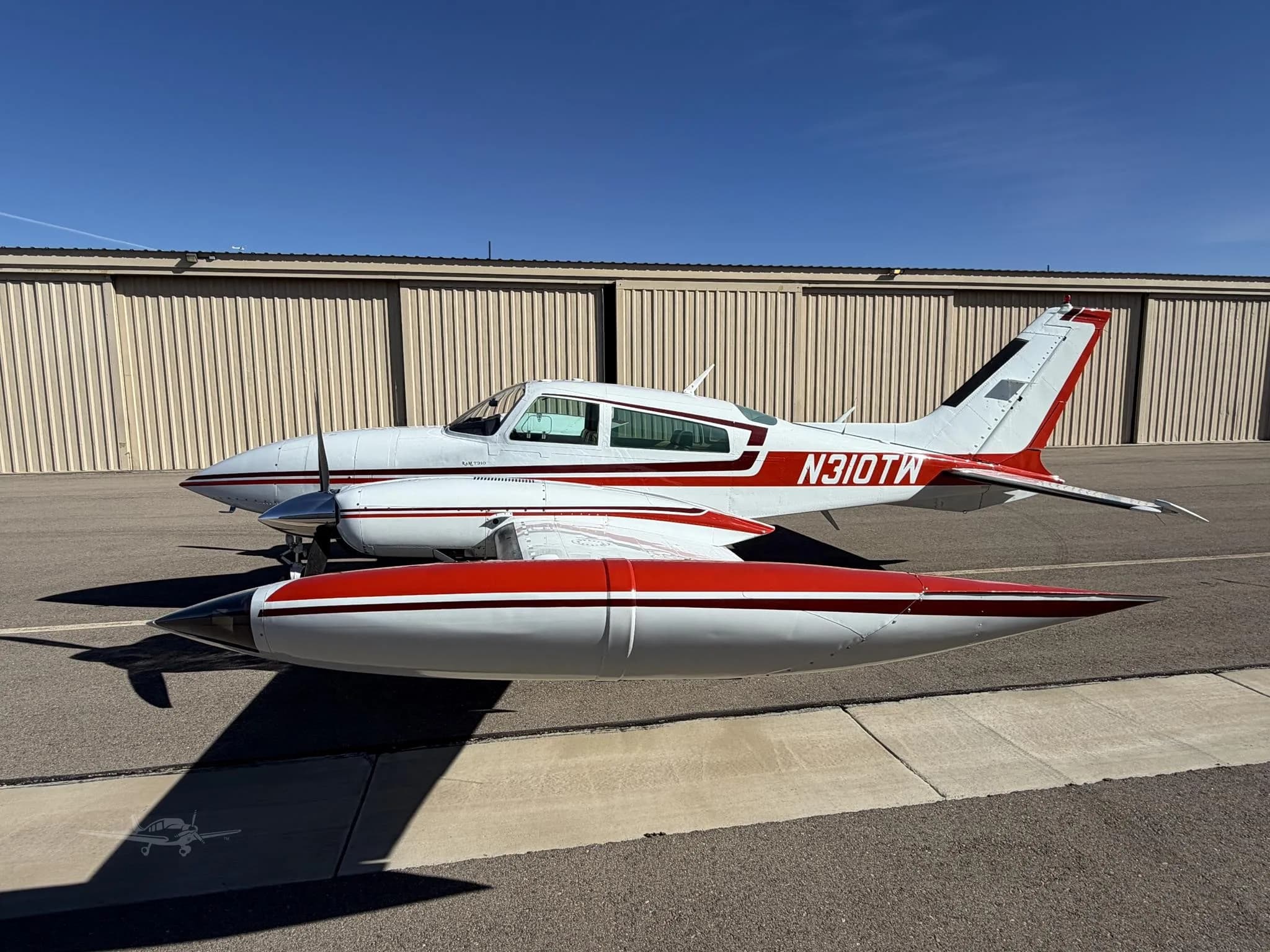 A white and red twin-engine Cessna 310 aircraft with registration N310TW is parked on an airfield in front of a hangar.