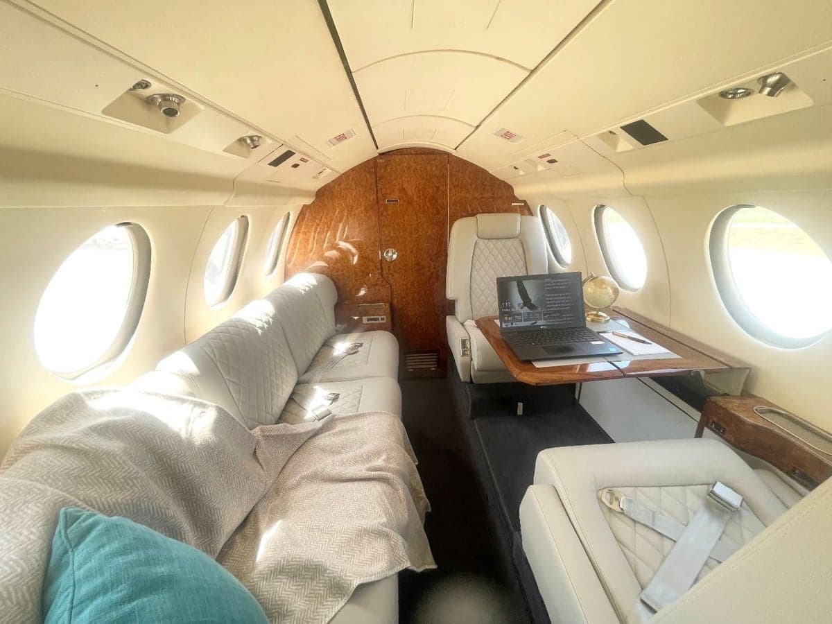 Aircraft interior with white leather seats and wood trim tables.