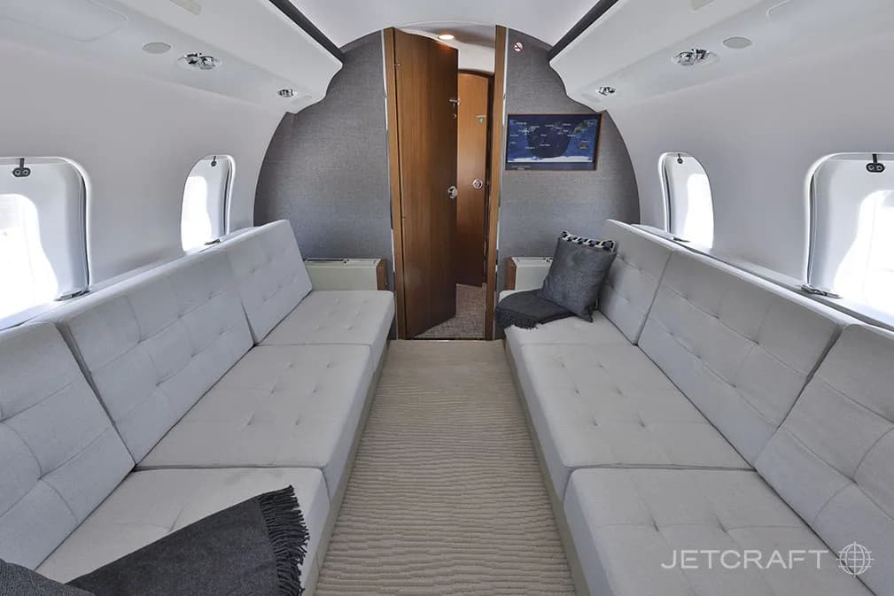 A view of the aft cabin of a Bombardier Global 6000, showing two light grey fabric sofas and an open door leading to another compartment.