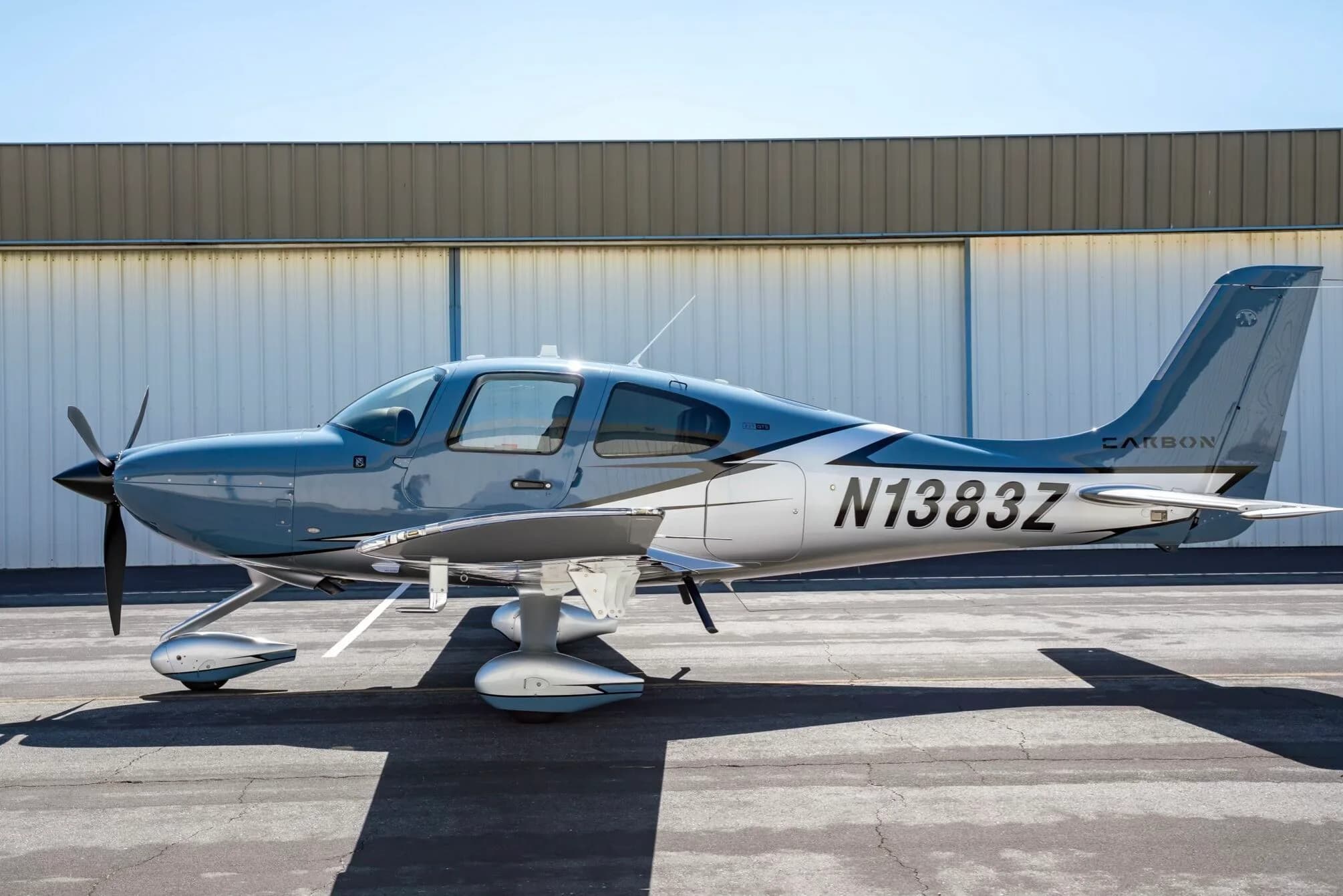 A light blue and silver Cirrus SR22T aircraft, N1383Z, parked on an tarmac in front of a hangar.
