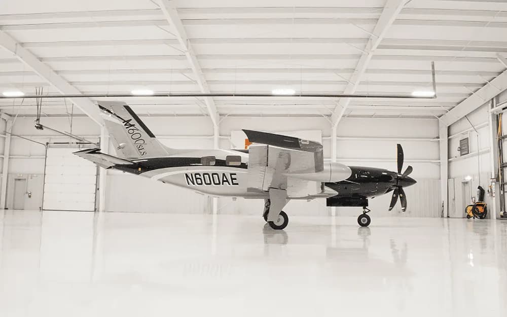 A Piper M600/SLS aircraft with registration N600AE, featuring a black and silver paint scheme, parked inside a brightly lit hangar.