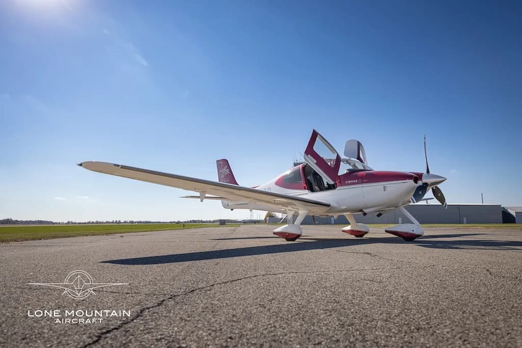 A white and maroon Cirrus SR22T aircraft with open doors parked on an airfield under a clear blue sky.