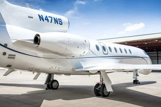 A rear-side view of a white Dassault Falcon 900EX business jet with blue stripes, registration N47NS, showing the engines and landing gear.