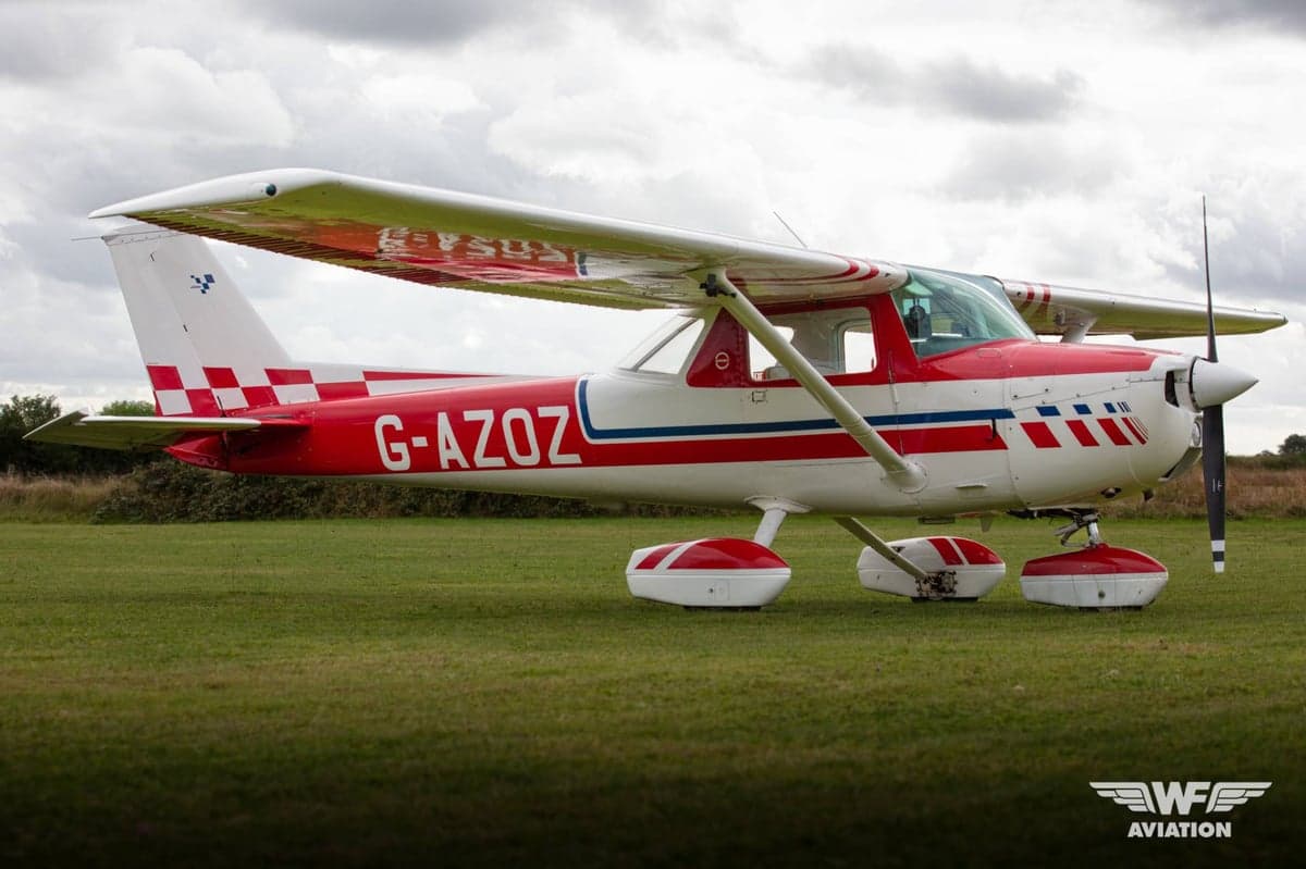 A red and white Cessna 150 Aerobat (G-AZOZ) with a checkered tail, parked on a grassy airfield.