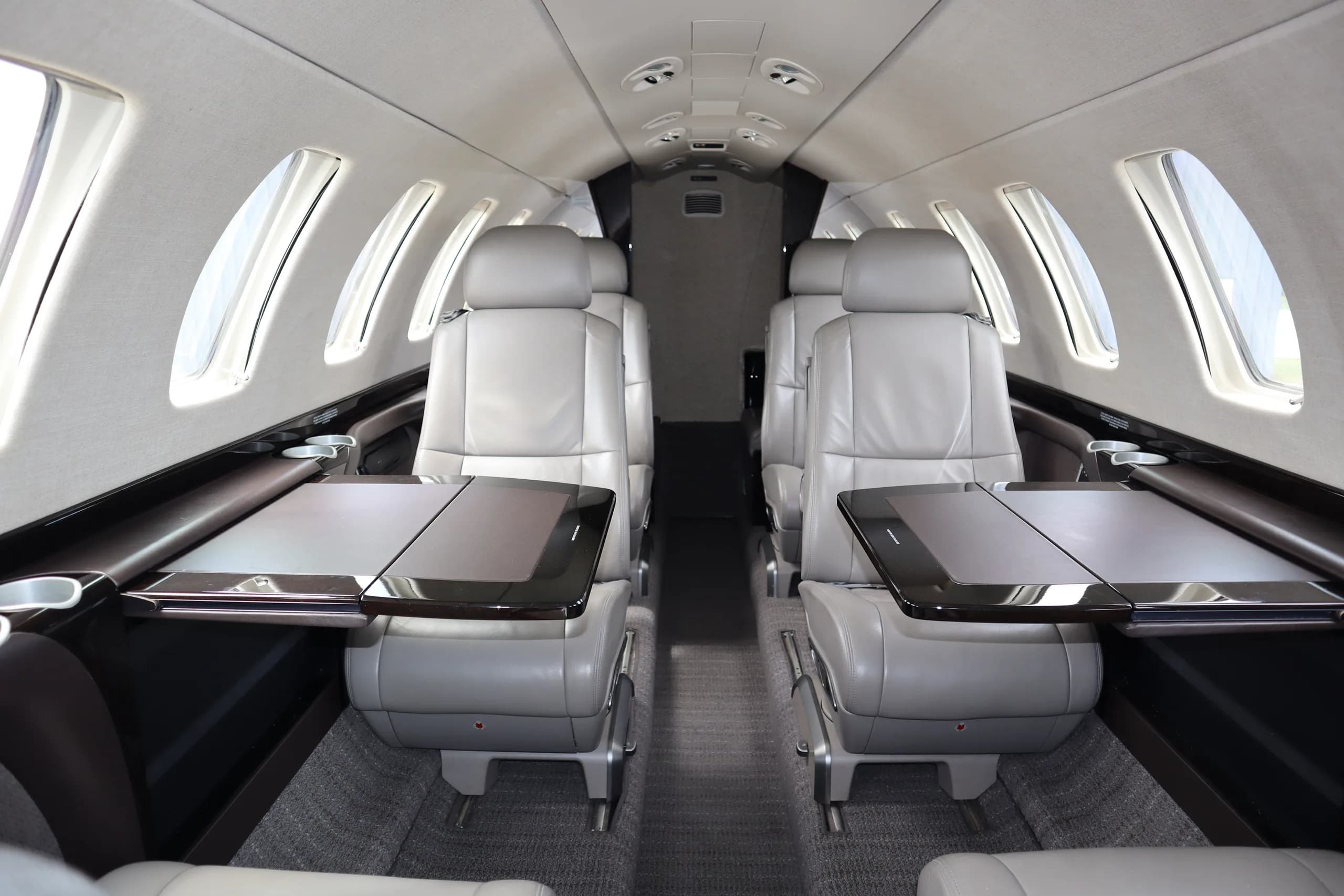 The interior cabin of a business jet, showing grey leather seats with fold-out tables, looking towards the rear.