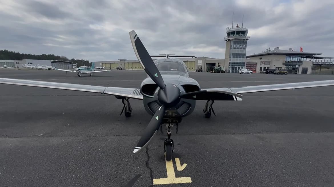 Front view of a gray Diamond DA50 RG aircraft parked on the tarmac with an airport control tower in the background.
