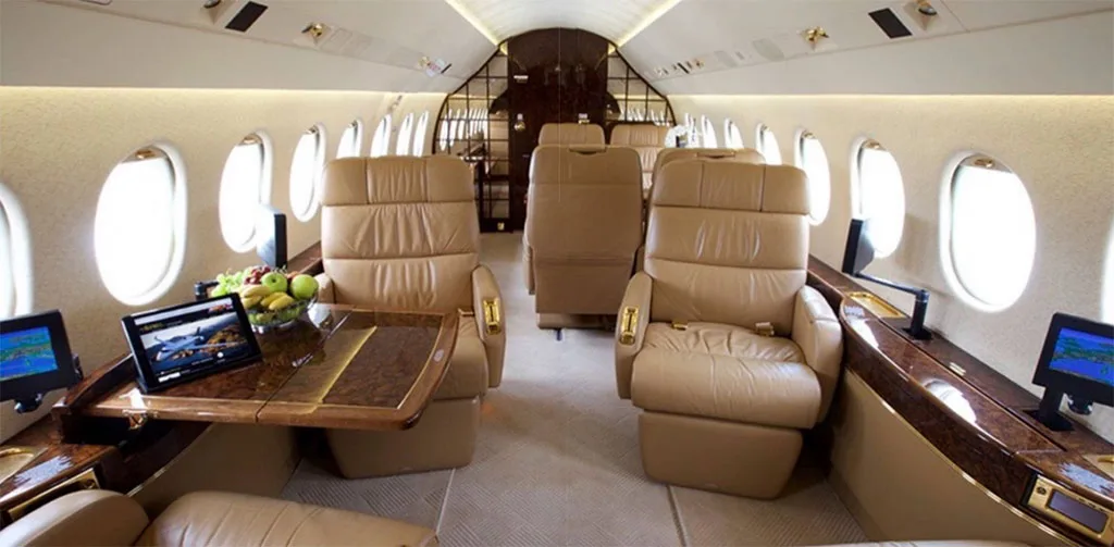The luxurious interior of a private jet cabin with tan leather seats, a polished wood table, and fresh fruit.