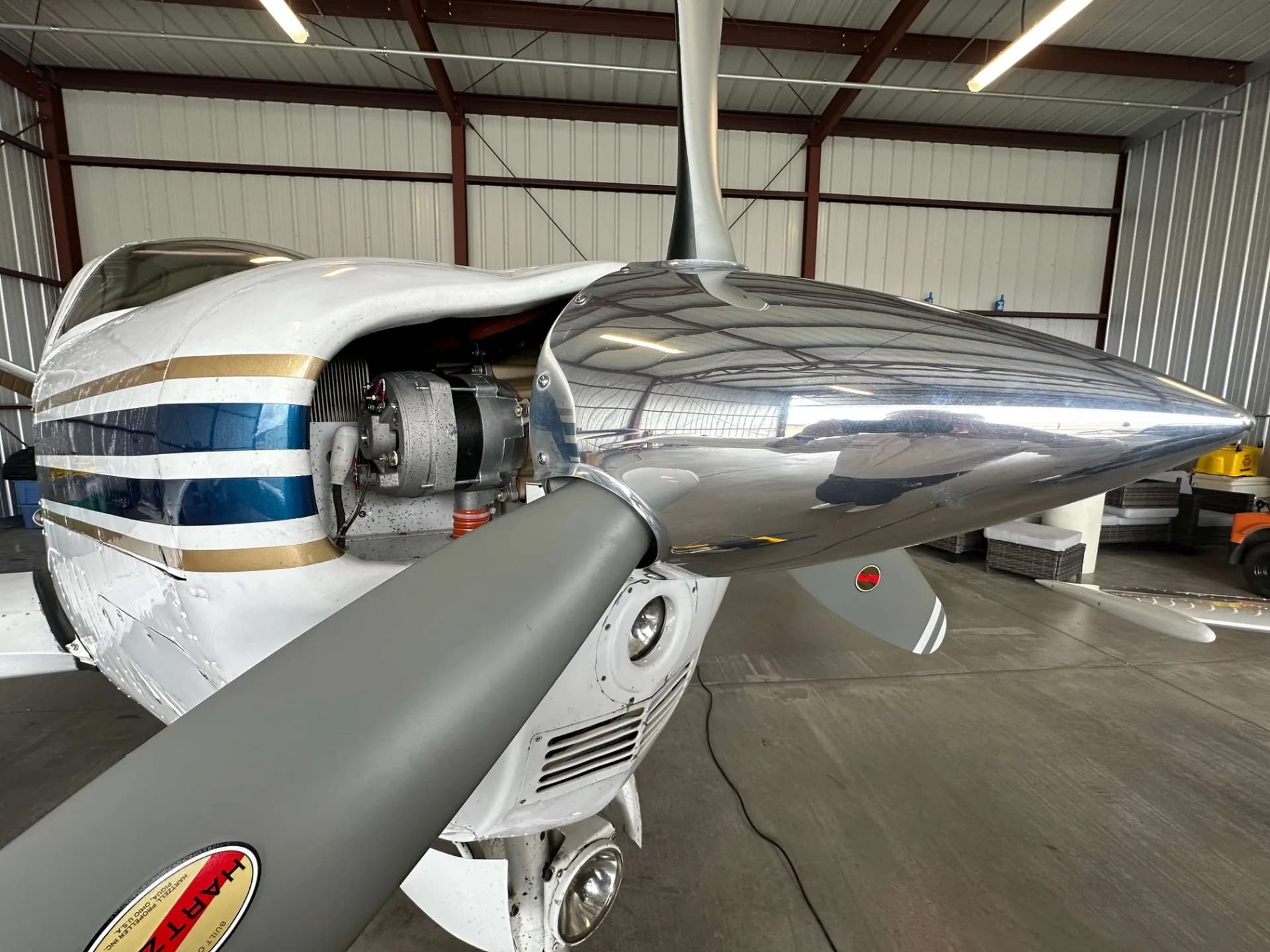 A close-up view of the polished propeller spinner and engine cowling of a white, blue, and gold Beechcraft Bonanza in a hangar.