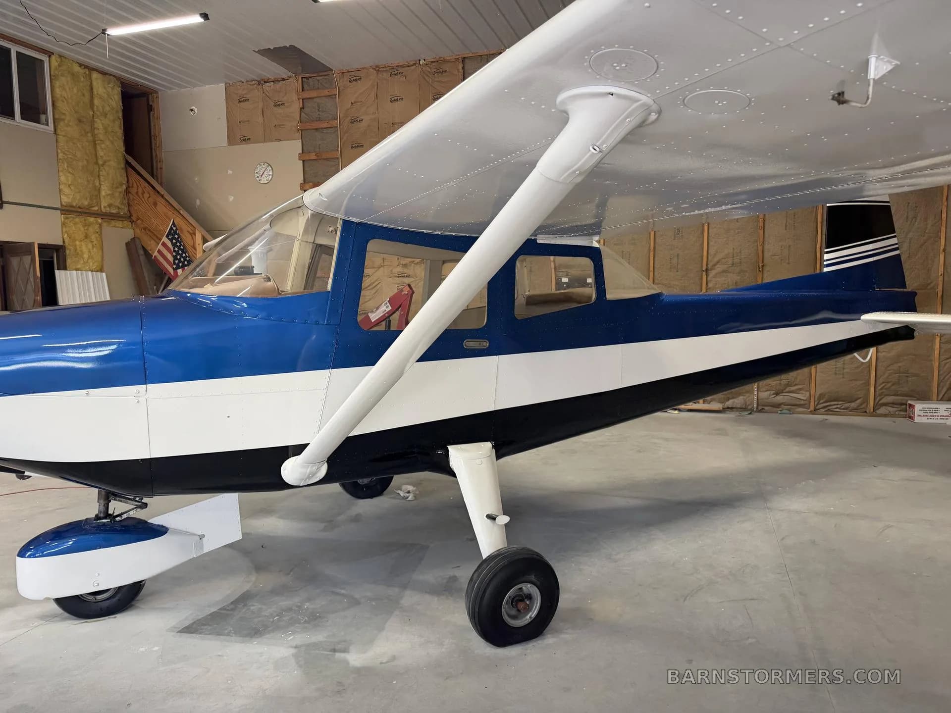 A blue and white high-wing aircraft, likely a Cessna, is parked in a hangar, viewed from the left side.