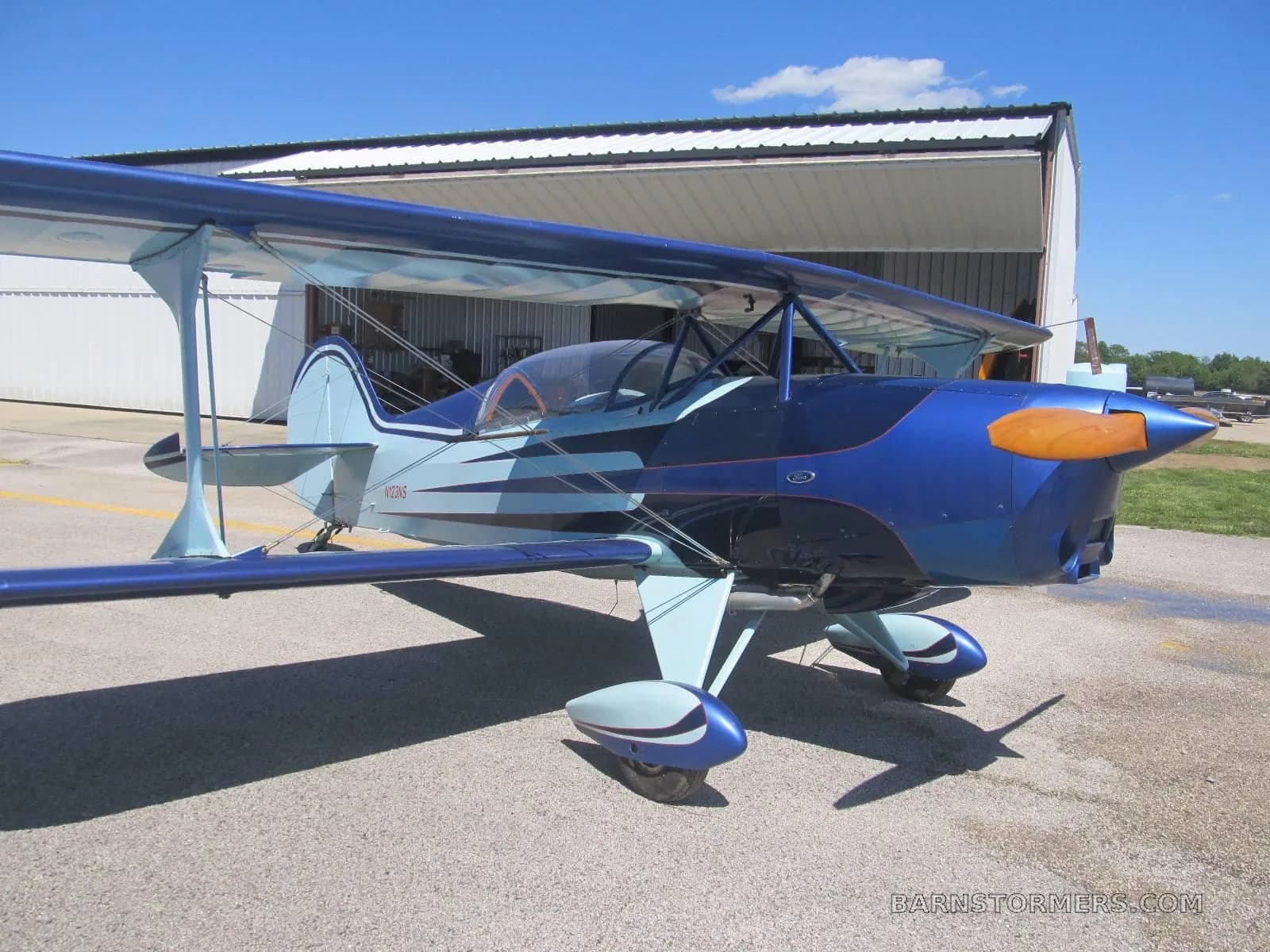 A blue and light blue biplane with an orange propeller spinner is parked on an asphalt apron in front of an open hangar.