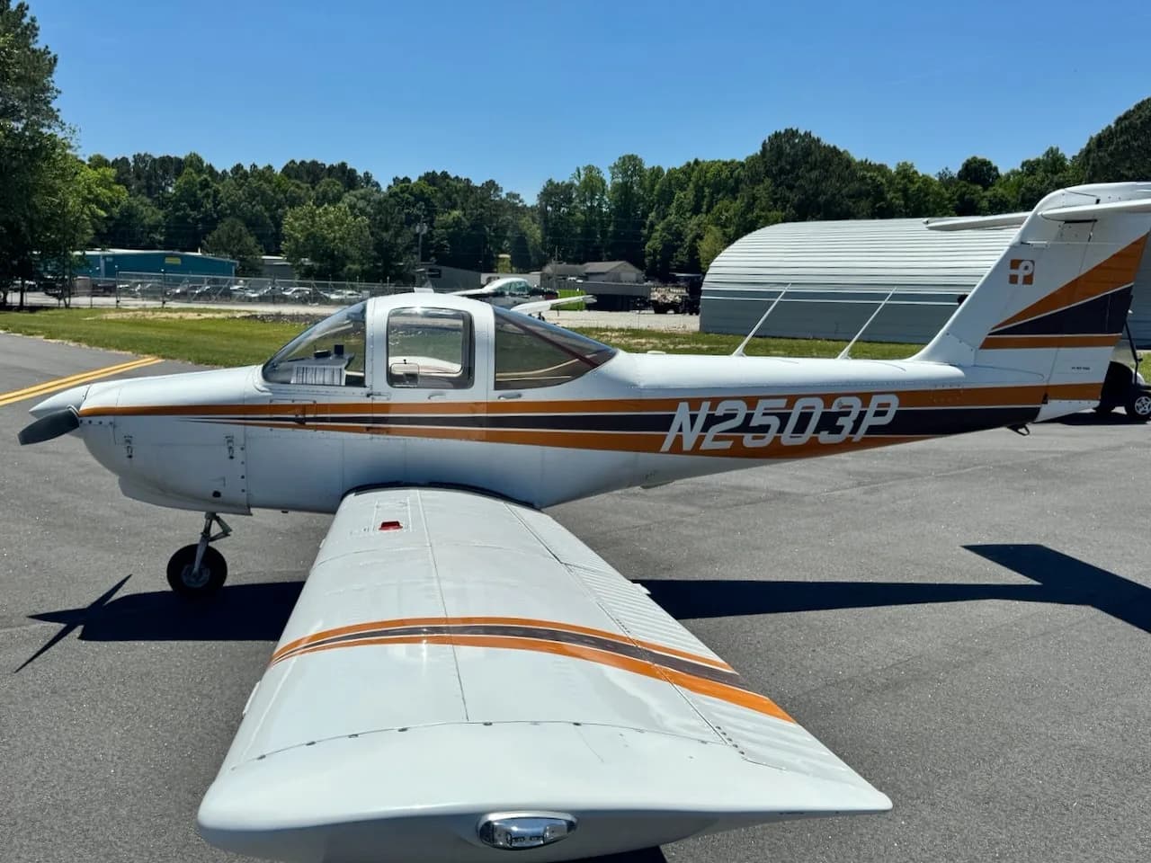 A white Piper PA-38 Tomahawk aircraft with orange and black stripes, seen from above the right wing on an airfield.