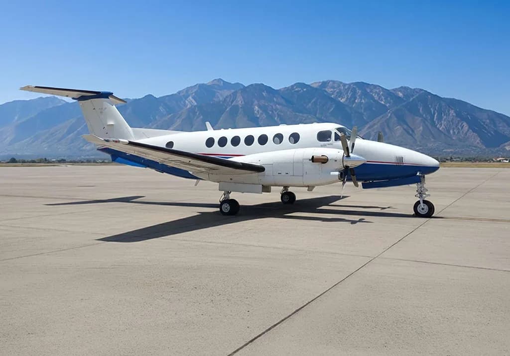A white and blue turboprop aircraft, likely a King Air, parked on an asphalt runway with mountains in the background under a clear blue sky.