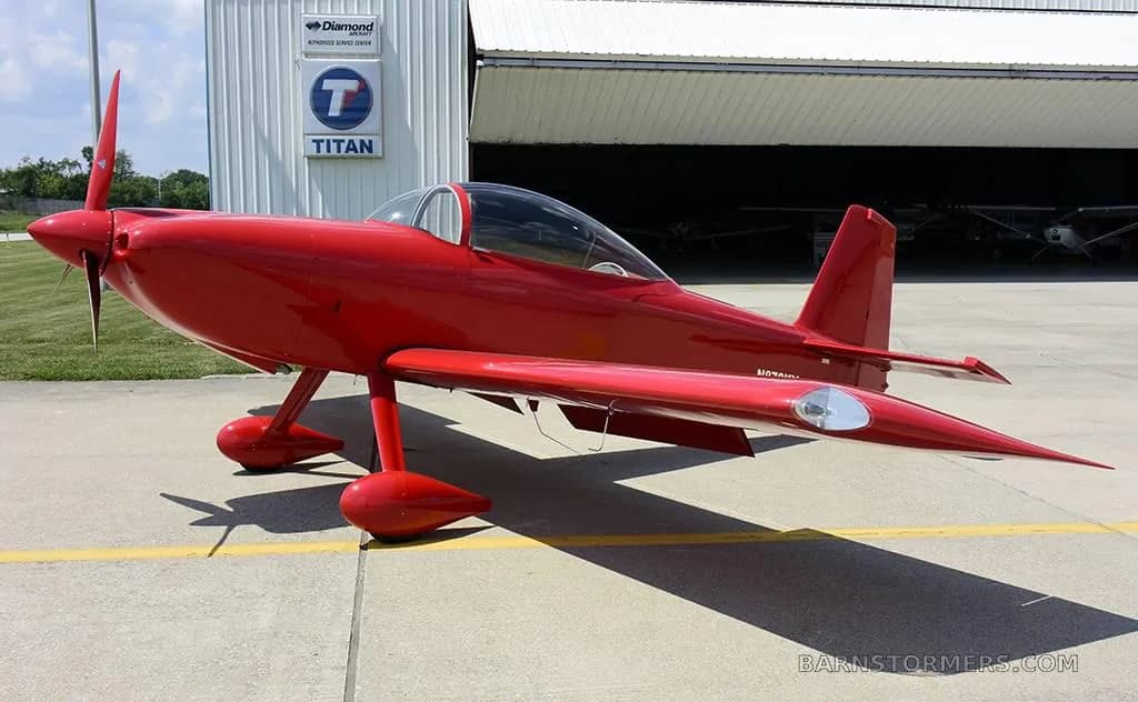 A red single-engine aircraft with a three-bladed propeller, parked on an airfield with a hangar in the background.