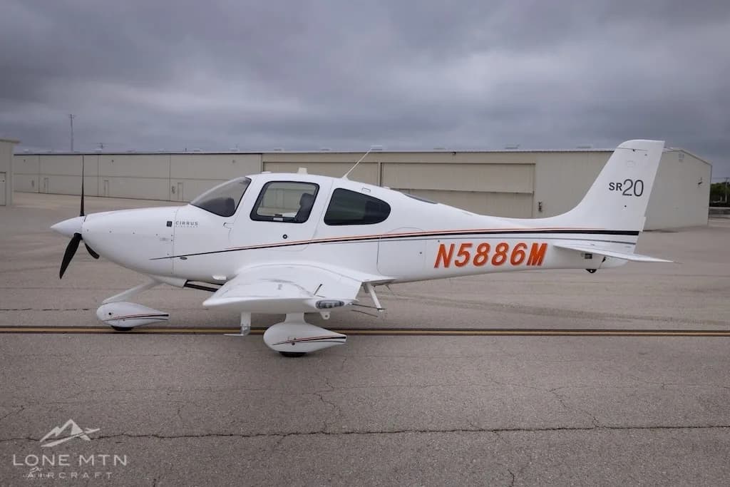 A full side profile of a white Cirrus SR20 aircraft with orange and black stripes, registration N5886M, parked on an airfield.