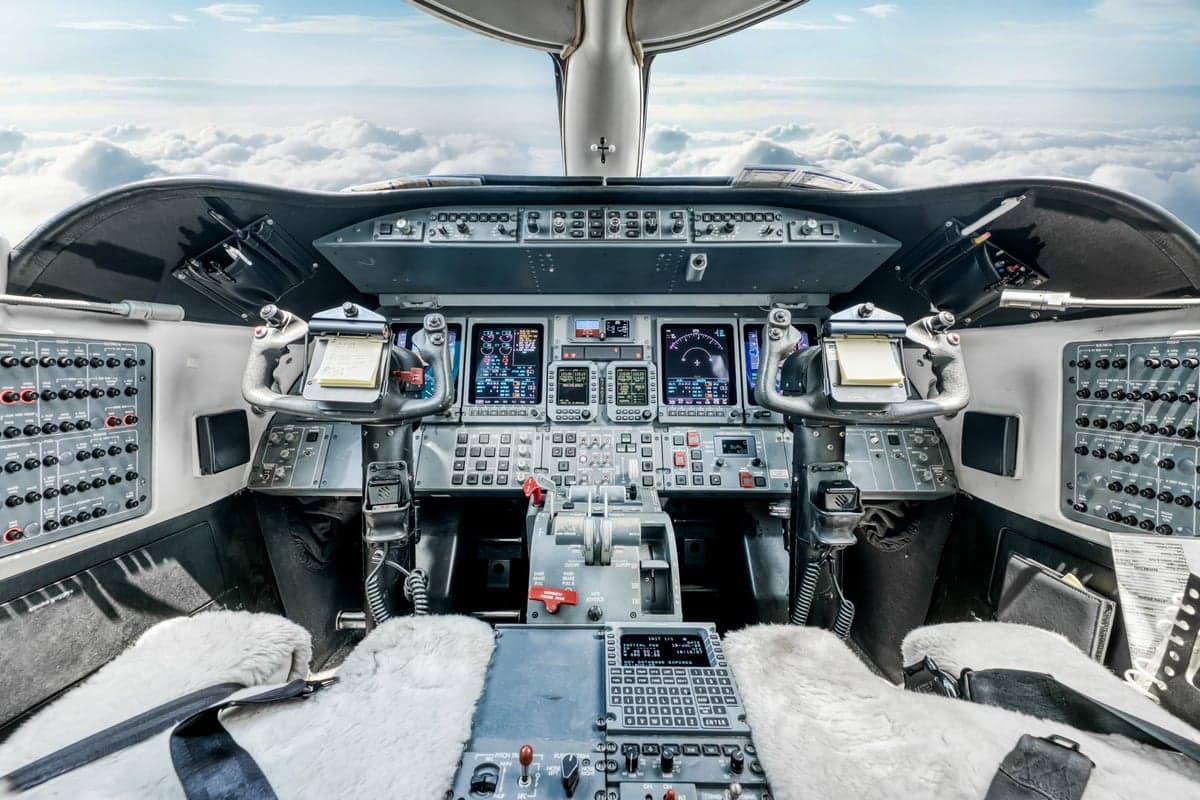 The modern cockpit of a private jet with multiple digital displays and controls, looking out over clouds.