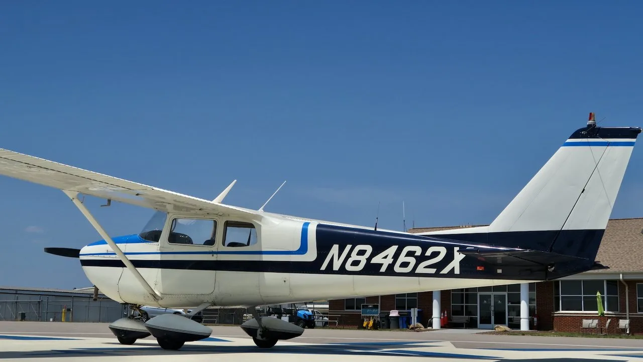 A side view of a white Cessna 172 with blue and black stripes, N8462X, parked in front of an airport building.