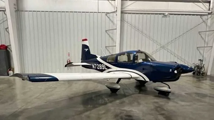 A blue and white Grumman American AA-5 Traveler sits inside a hangar.