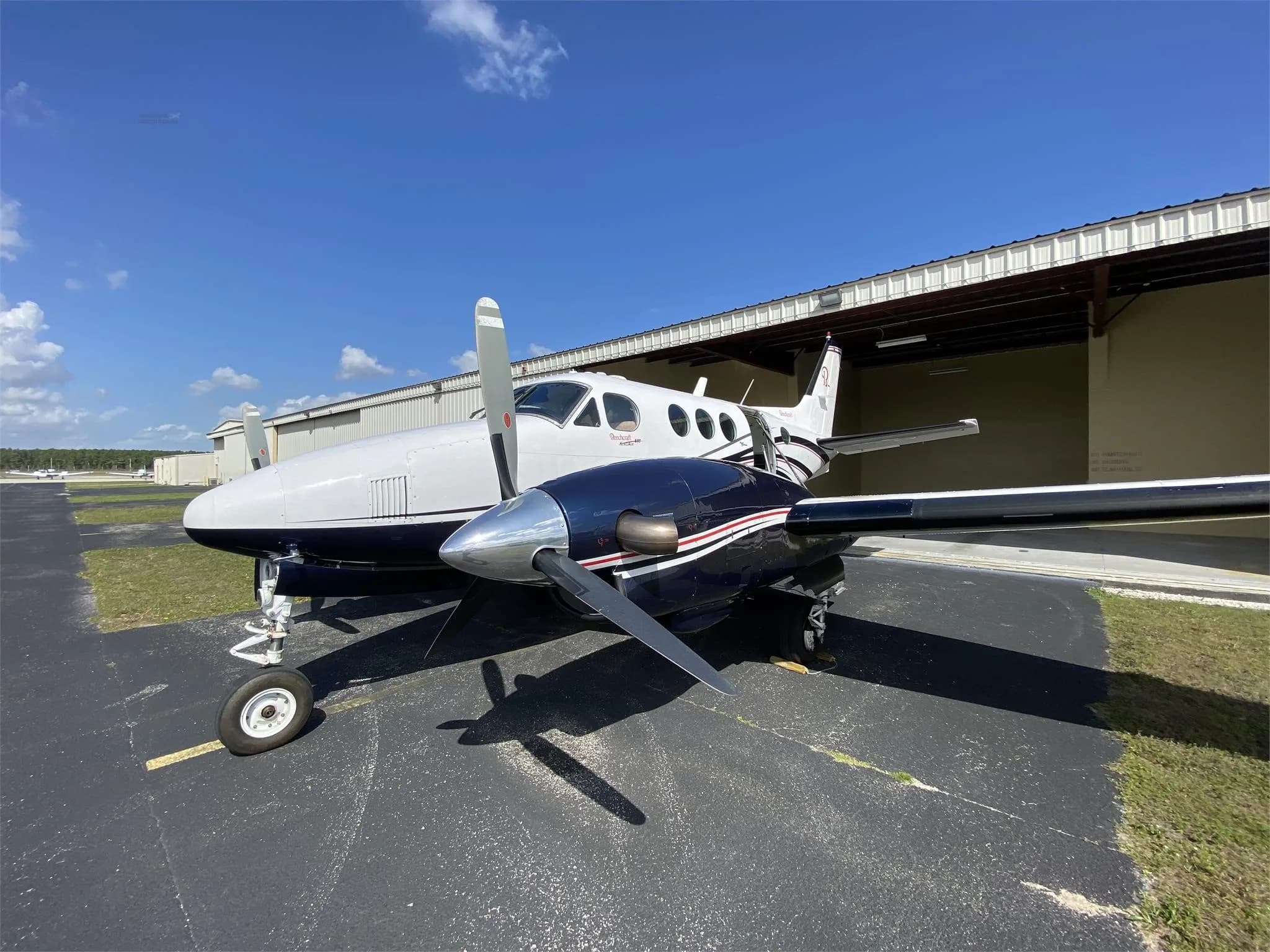 A white and dark blue twin-engine turboprop aircraft is parked on an asphalt tarmac next to a hangar under a clear blue sky.