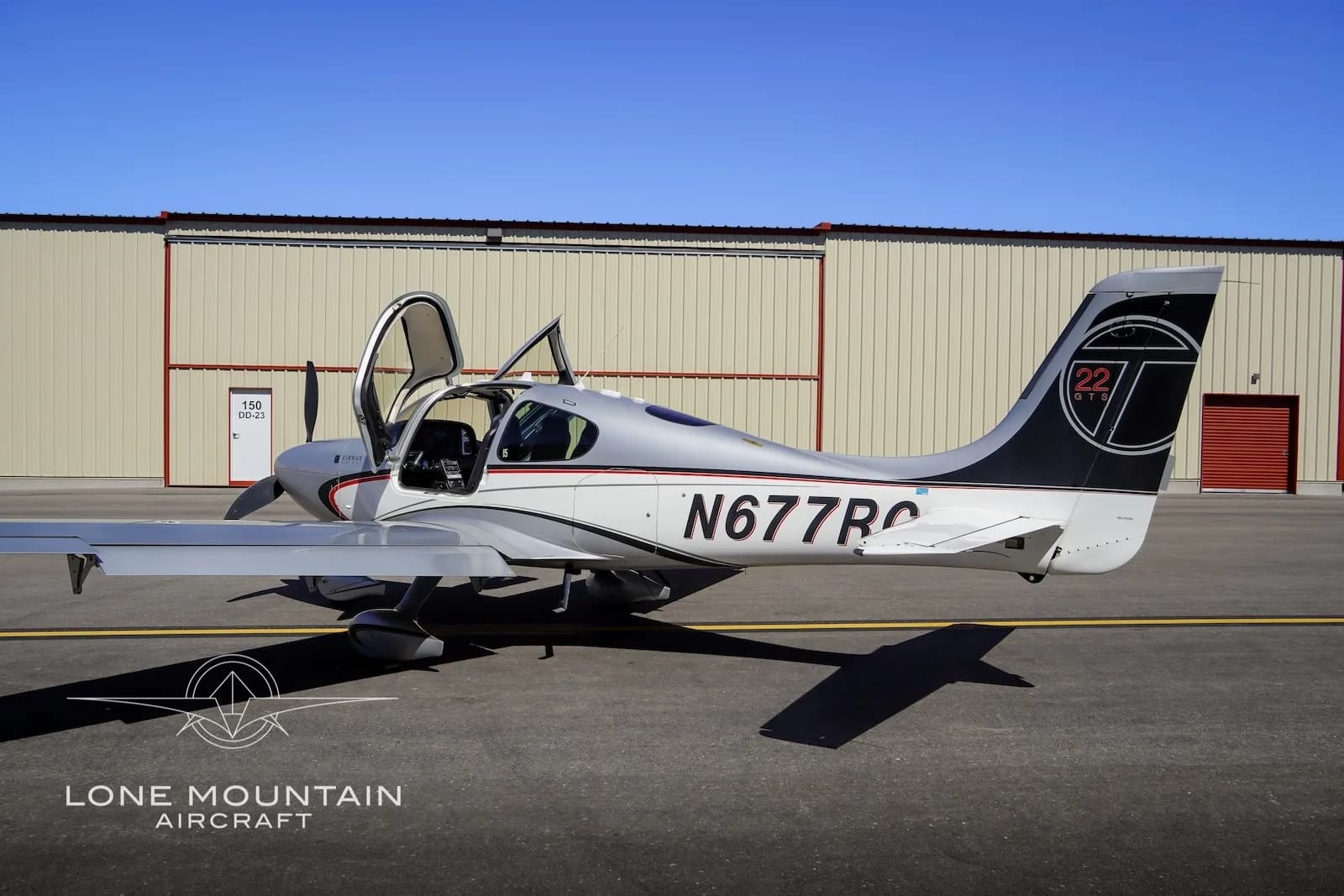 A silver Cirrus SR22 aircraft with red and black accents, registration N677RC, parked on an airfield with both doors open, viewed from the side.