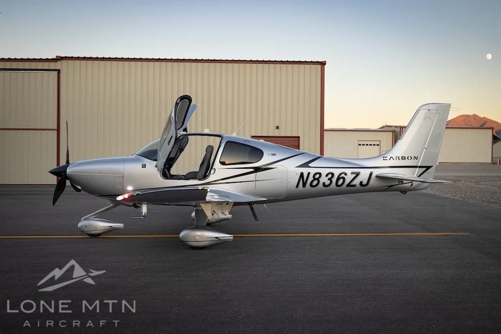 A silver Cirrus SR22T aircraft with registration N836ZJ is parked on an asphalt ramp with its doors open, showing its side profile against hangars and a distant moon.