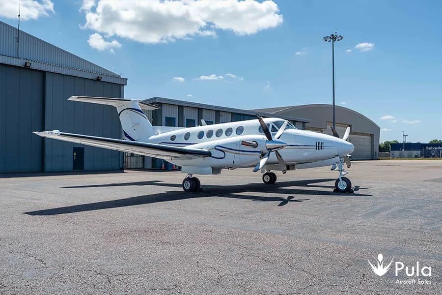 A white twin-engine turboprop aircraft with blue stripes is parked on an airport tarmac next to hangars under a blue sky.