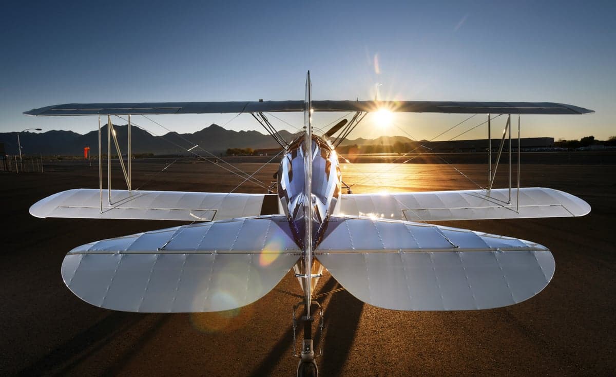 A biplane is viewed from the front, showing the wings and propeller, with the sun setting in the background.