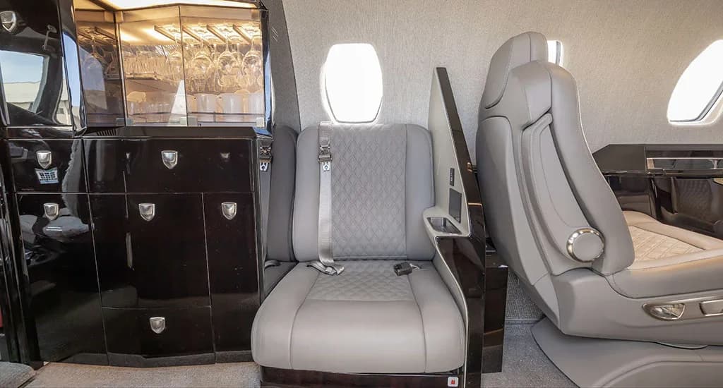 A close-up of the private jet's interior showing a dark wood galley with glassware and a light gray quilted leather seat.