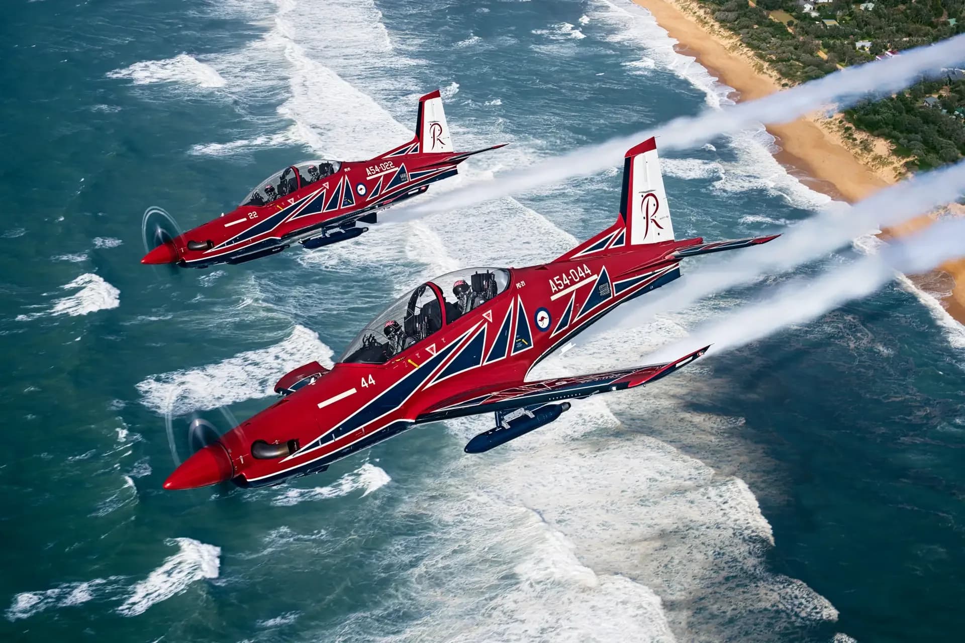 Two red and white Pilatus PC-21 aircraft flying in formation over a beach and ocean.